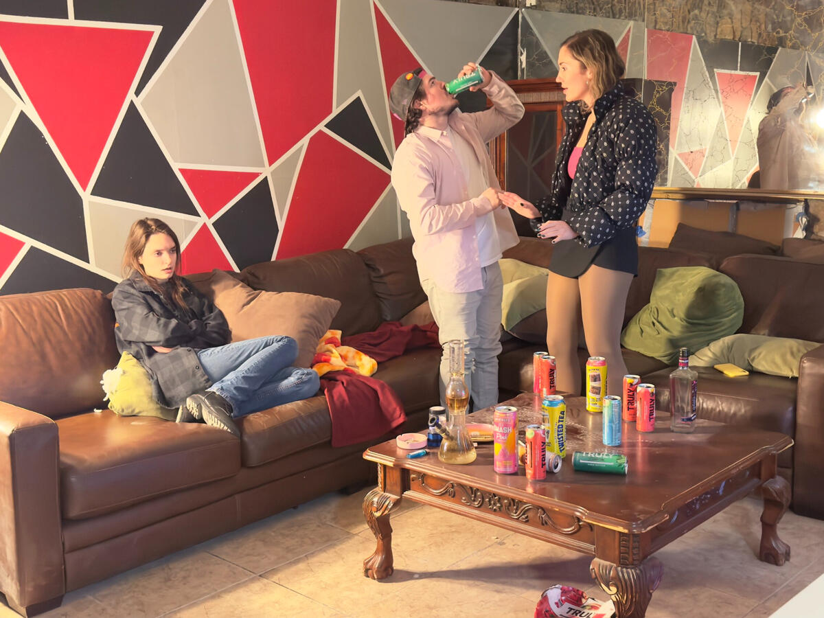 Self Preservation by Linsey Lewis (2024) A girl sitting on the couch surrounded by drug paraphernalia and empty beer cans with a boy and girl arguing to the right, with the boy drinking a beer.